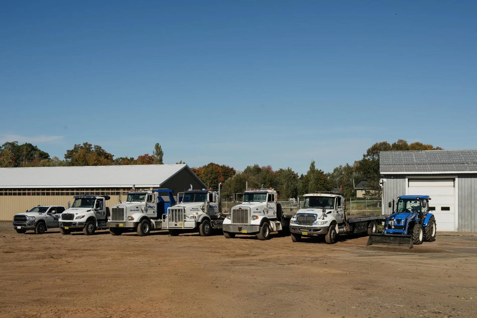 The whole Milne's fleet lined up in the yard: a pickup, white tow trucks and flatbeds, and a blue tractor.